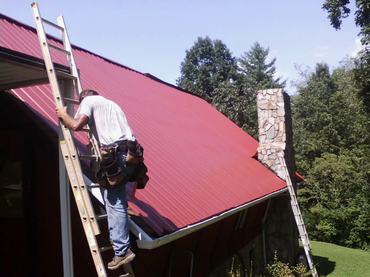 Roofer on a ladder beside a red metal roof and stone chimney — Soffit & Fascia Repair by Roofing Brothers in Timberlake